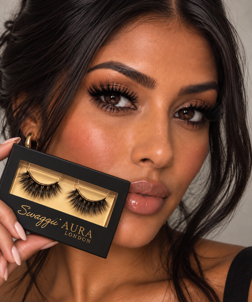 A close-up of a woman holding a black Swaggii Aura London false eyelash pack next to her face, showcasing the black lashes inside the clear window of the packaging.